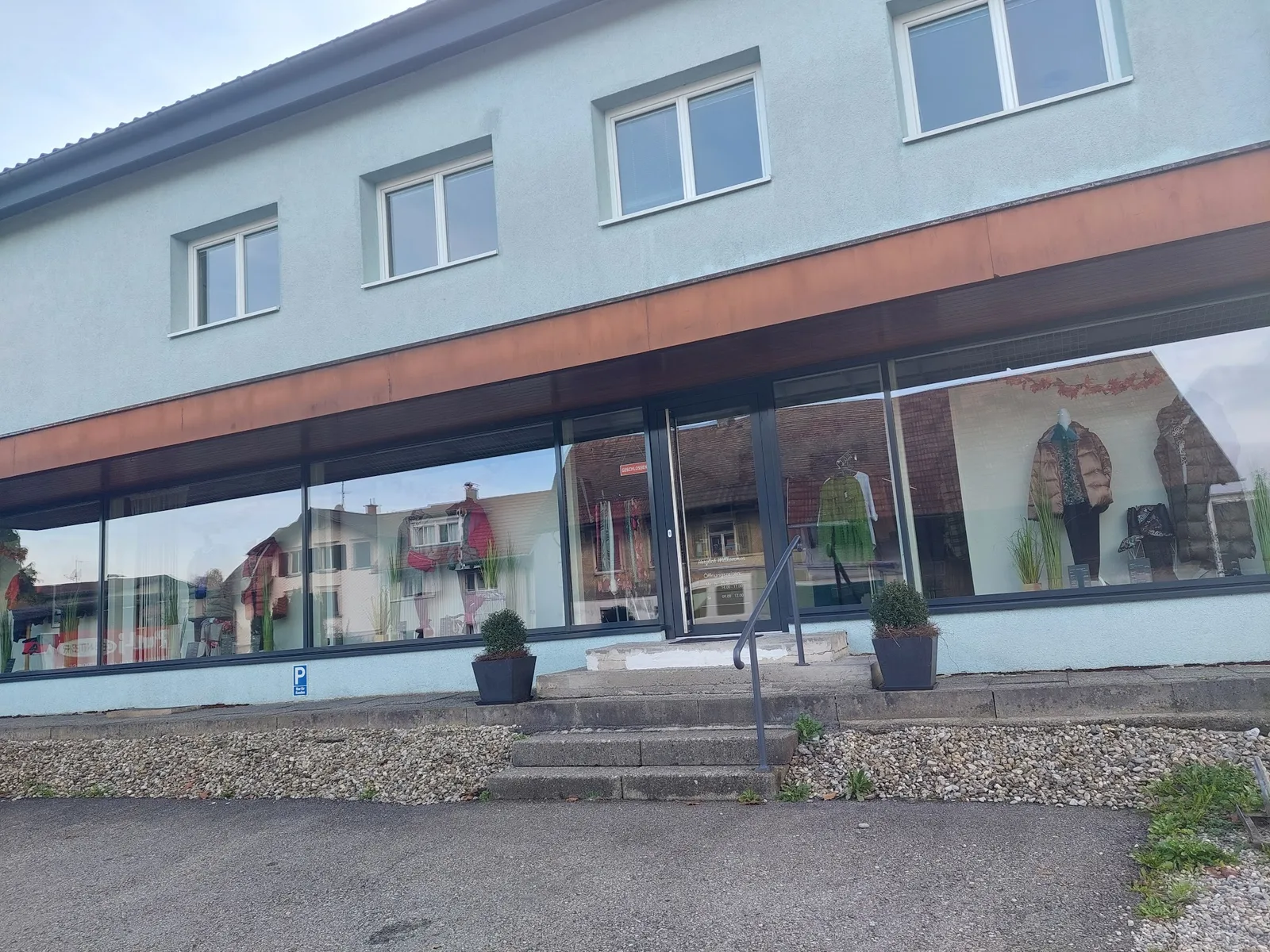 Außenansicht des Geschäfts: helles Gebäude mit durchgehender Schaufensterfront, Eingangstür mit Stufen und Handlauf, davor ein Parkplatz